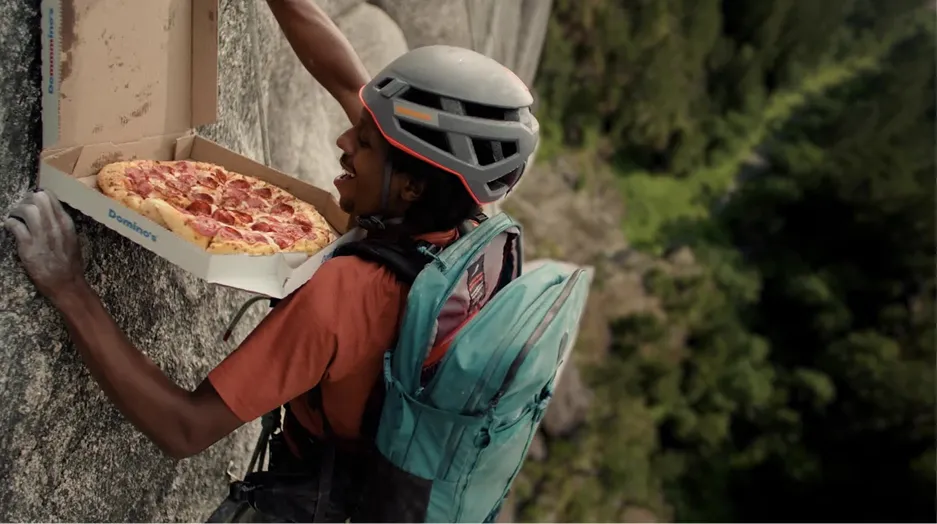 A rock climber opens a Domino's pizza box while hanging from a sheer cliff face